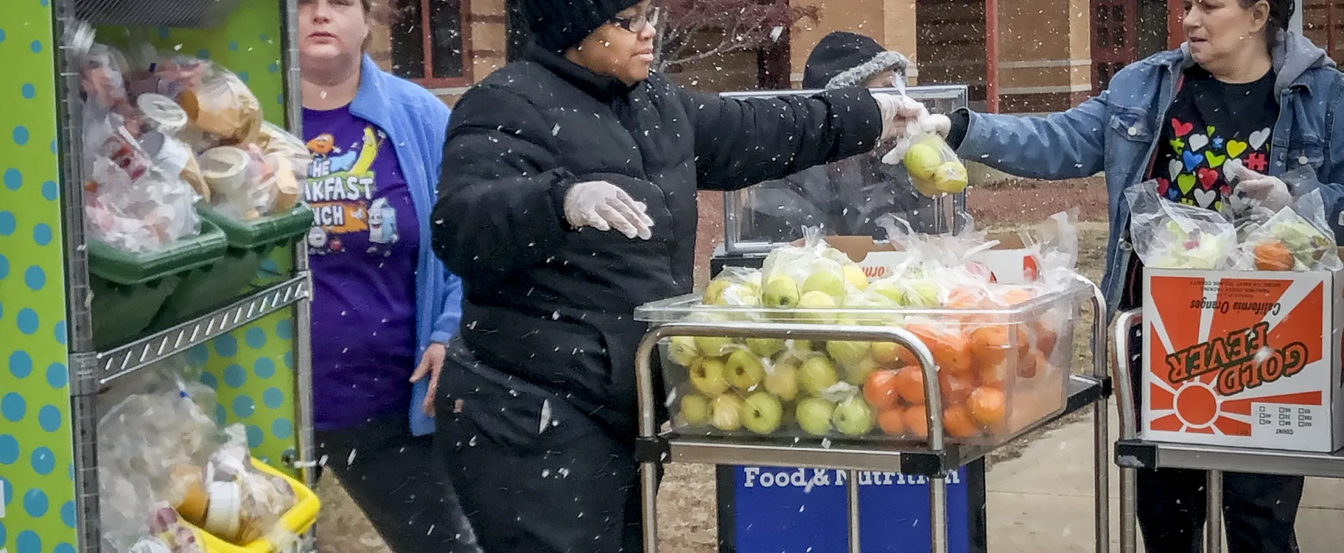 Three food service workers handle bags of fruit outside in the snow.