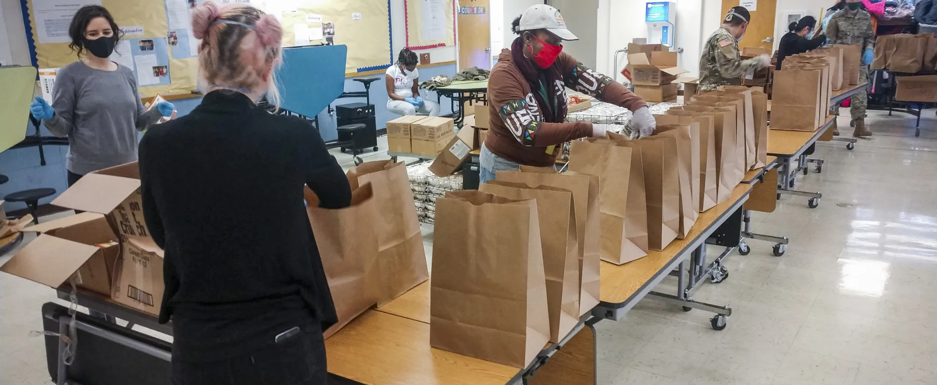 A group packs meals in paper bags on tables in a cafeteria.