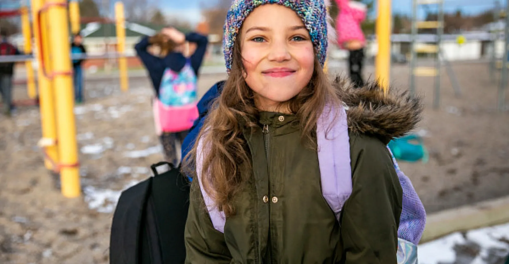 A girl in a beanie and coat smiles on her school playground.