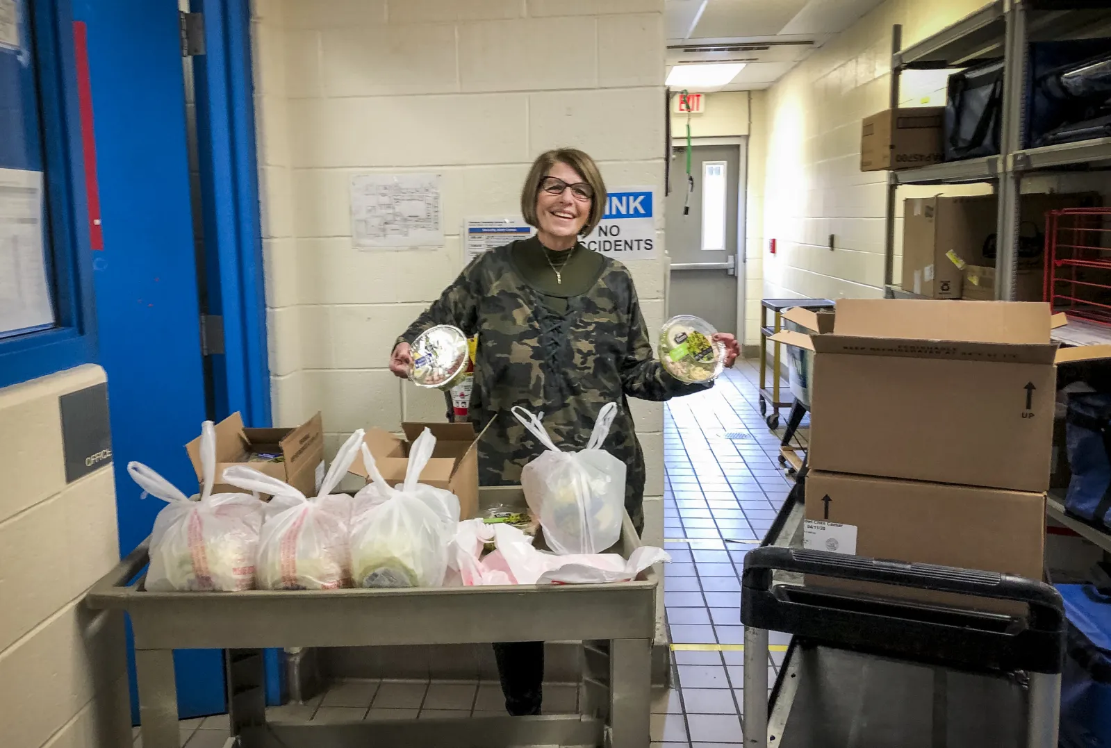 A woman holds up two donated, packaged salads behind a cart packed with meals for families need.