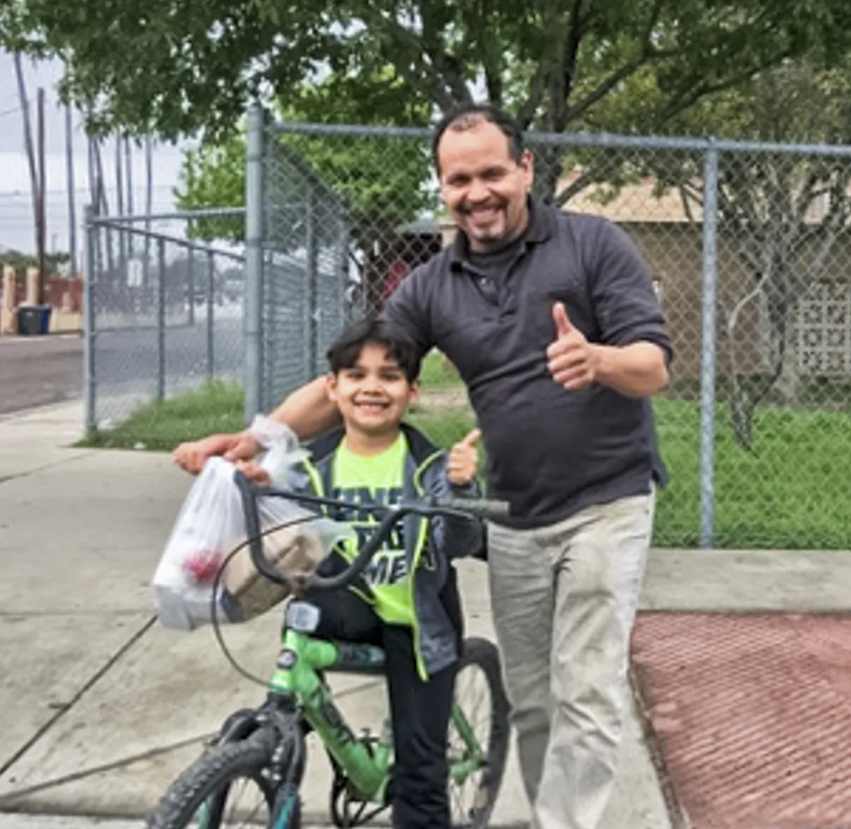 A father and son give thumbs up as they leave a meals site on their bike with a meal bag.