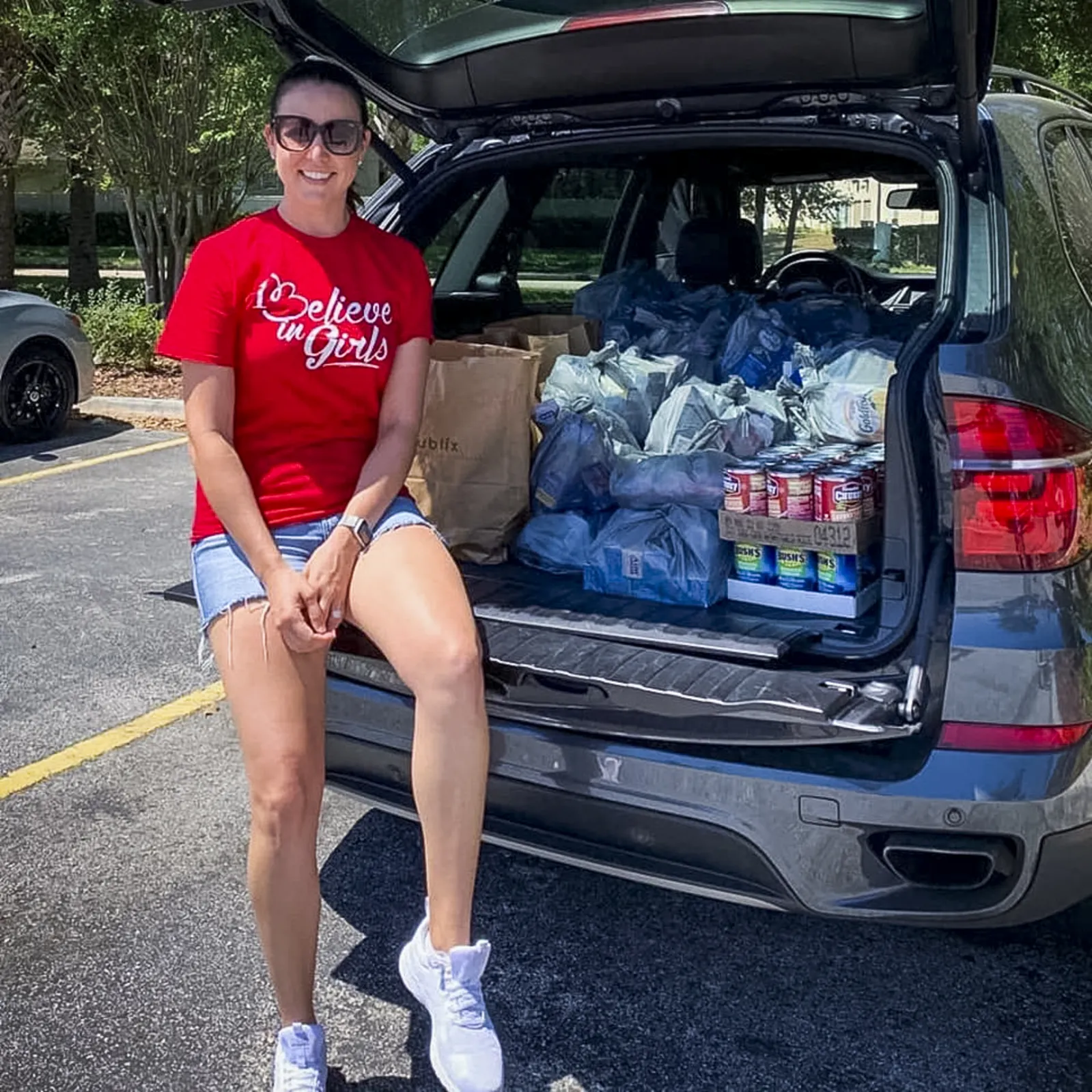 A woman sits in the back of her open SUV next to piles of food.