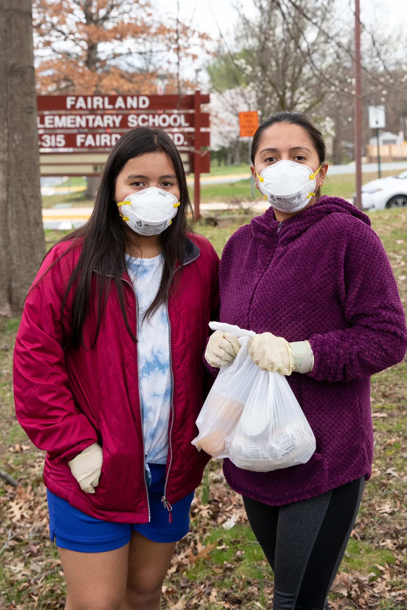 A Latina mother and daughter wearing masks and gloves hold a bag of food supplies on a fall day.