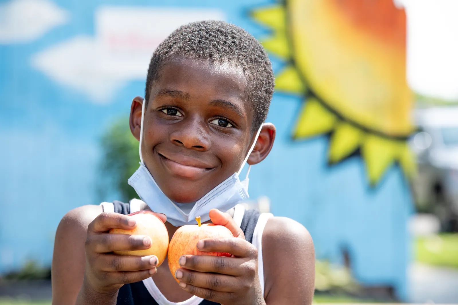 Black kid with mask under the chin holding two apples and happy