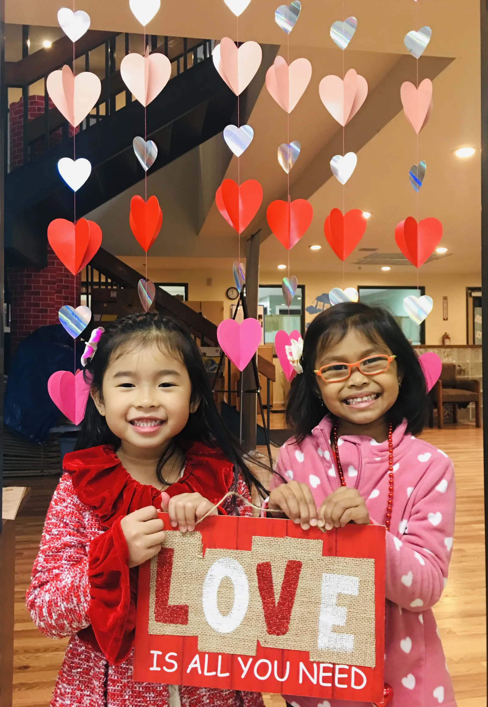 Two girls holding a sign that says love is all you need. They have hearts in the background