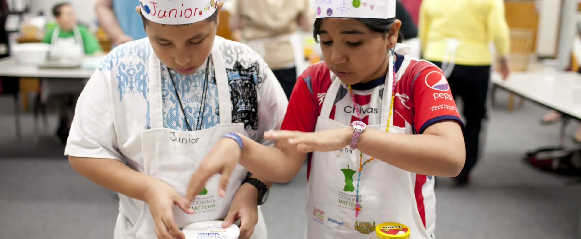 Two kids wearing aprons and chef hats looking at baking ingredients