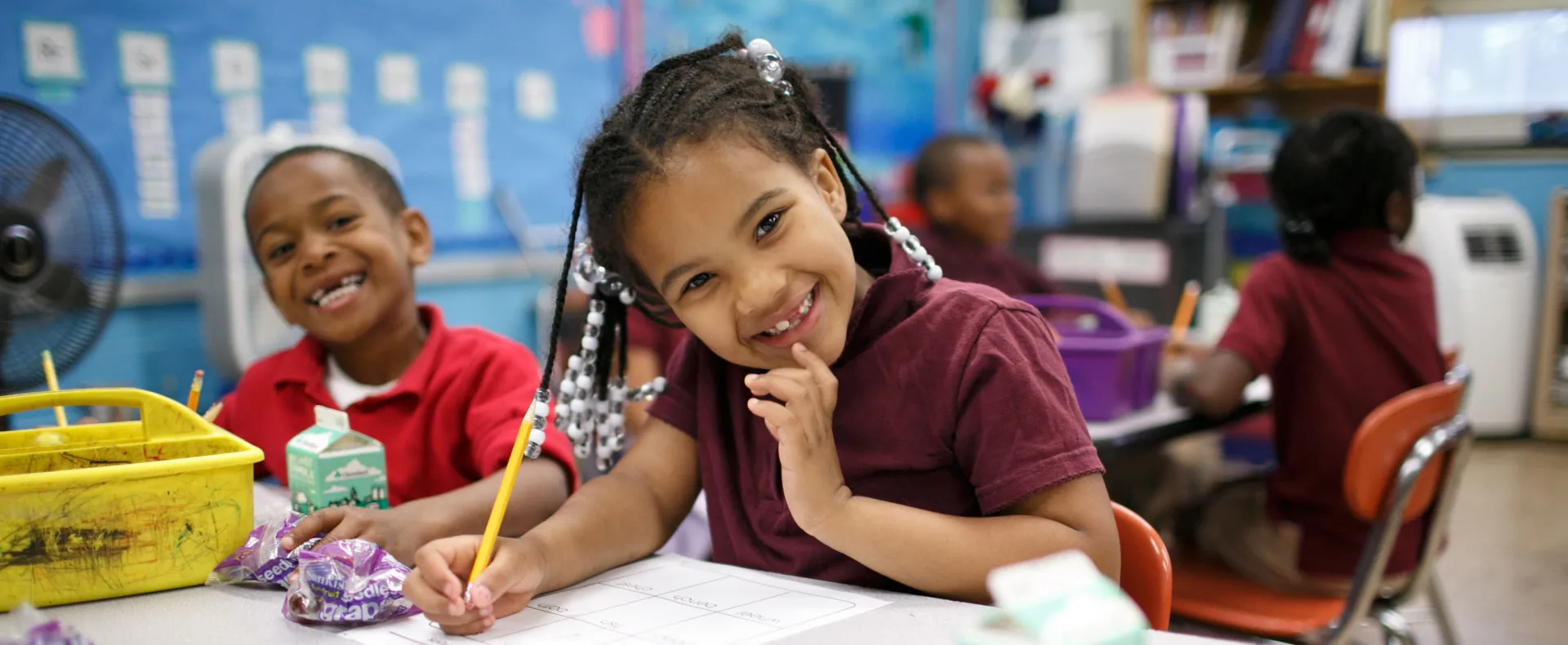 Children sitting at school table working on worksheets with breakfast on the table