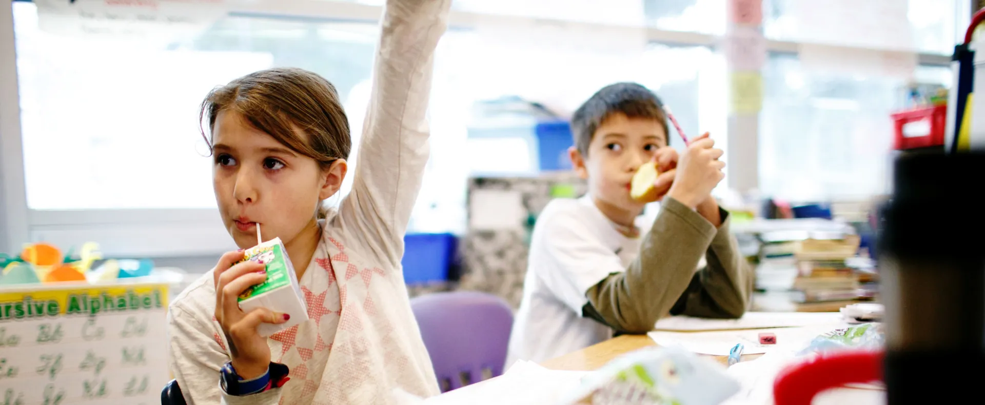 Children eating breakfast at desks and raising their hands