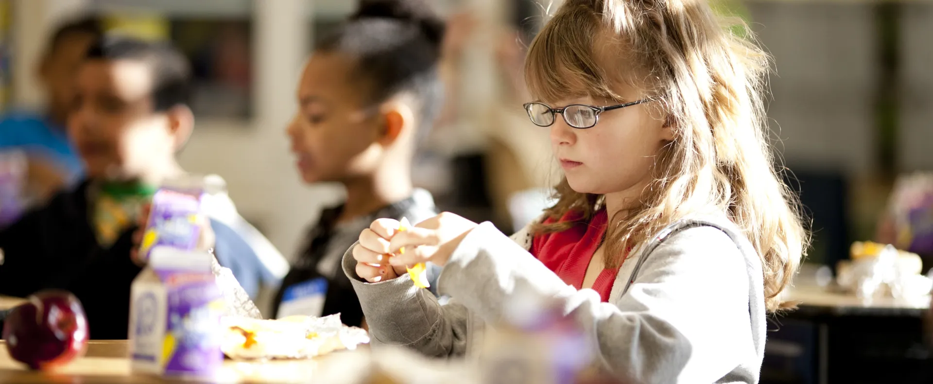 A little girl with glasses eats breakfast in her classroom.