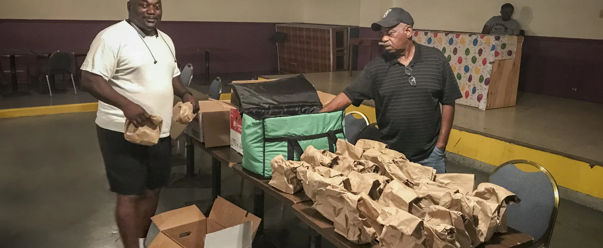 Two men pack paper meals bags at a table in a low-lit room.