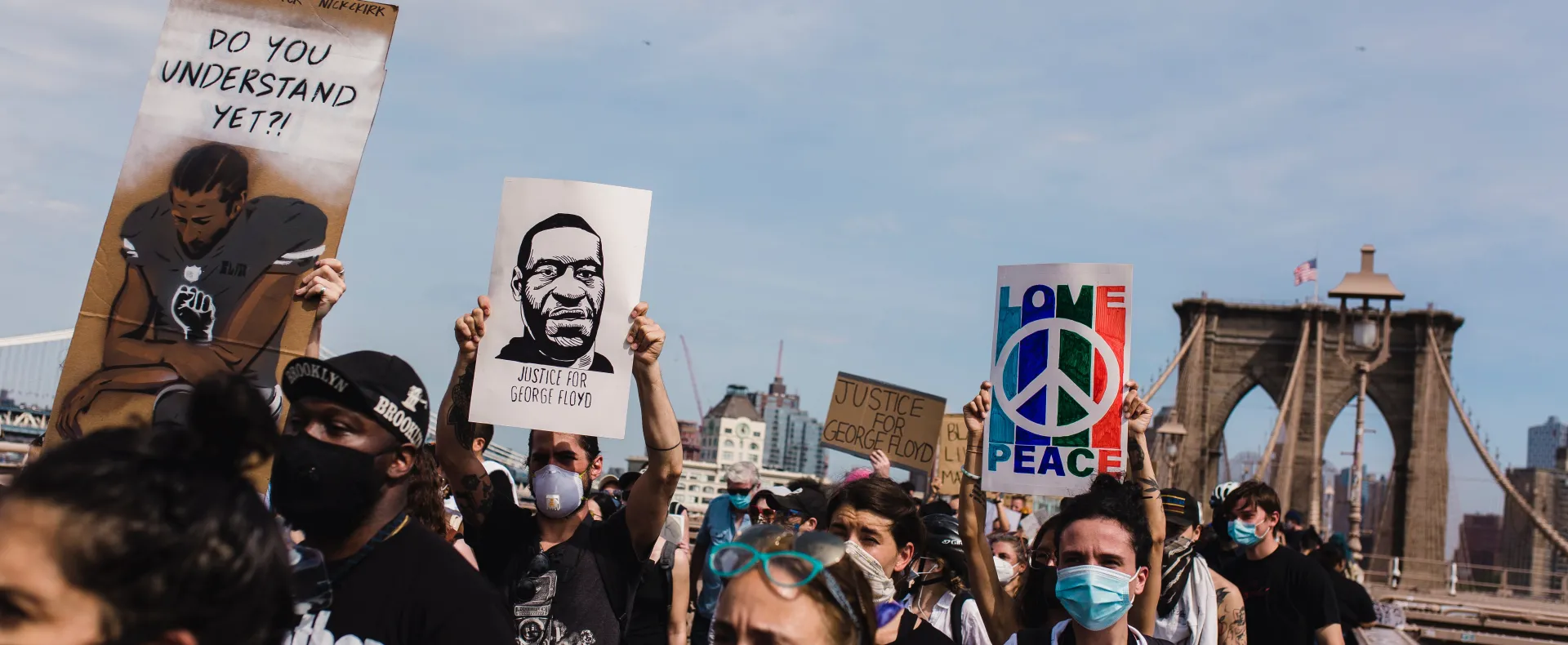 Racial justice protestors in coronavirus masks cross the Brooklyn bridge carrying signs remembering George Floyd.