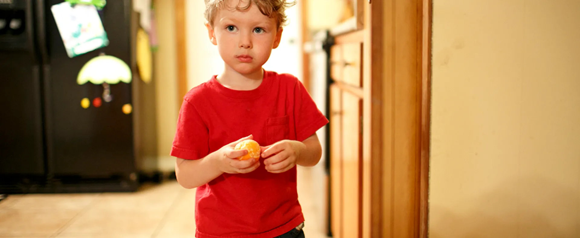 A little boy with a sad look on his face stands in his kitchen