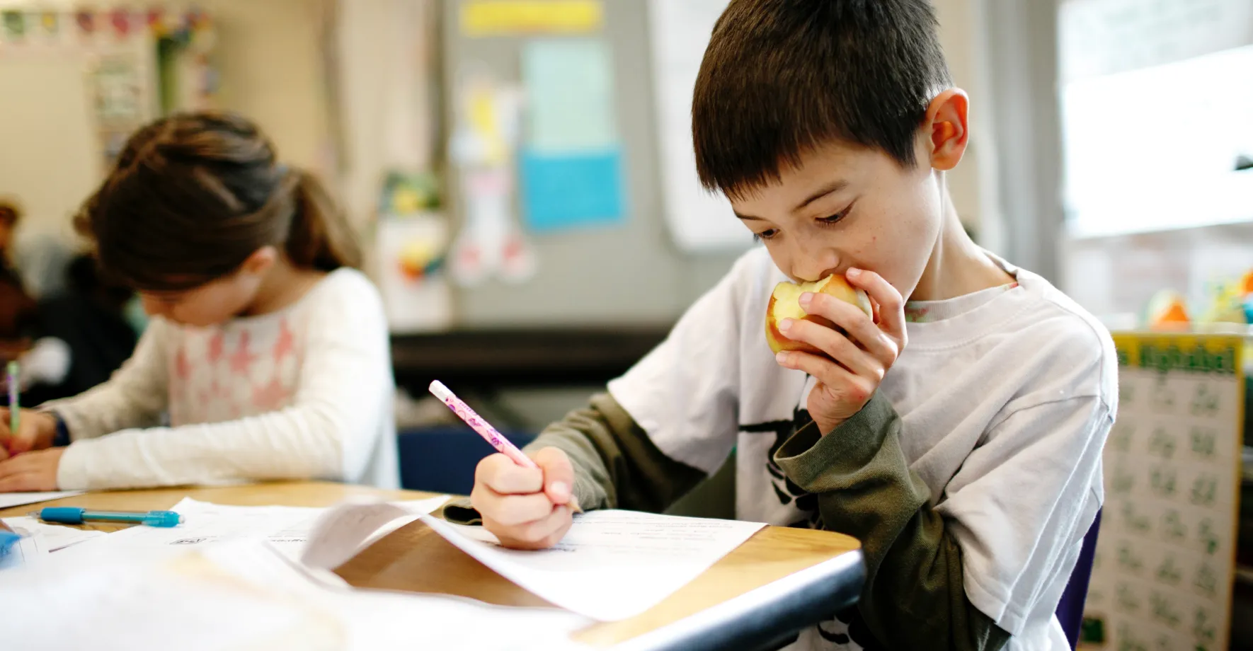 Little boy eating an apple while working on schoolwork in the classroom.
