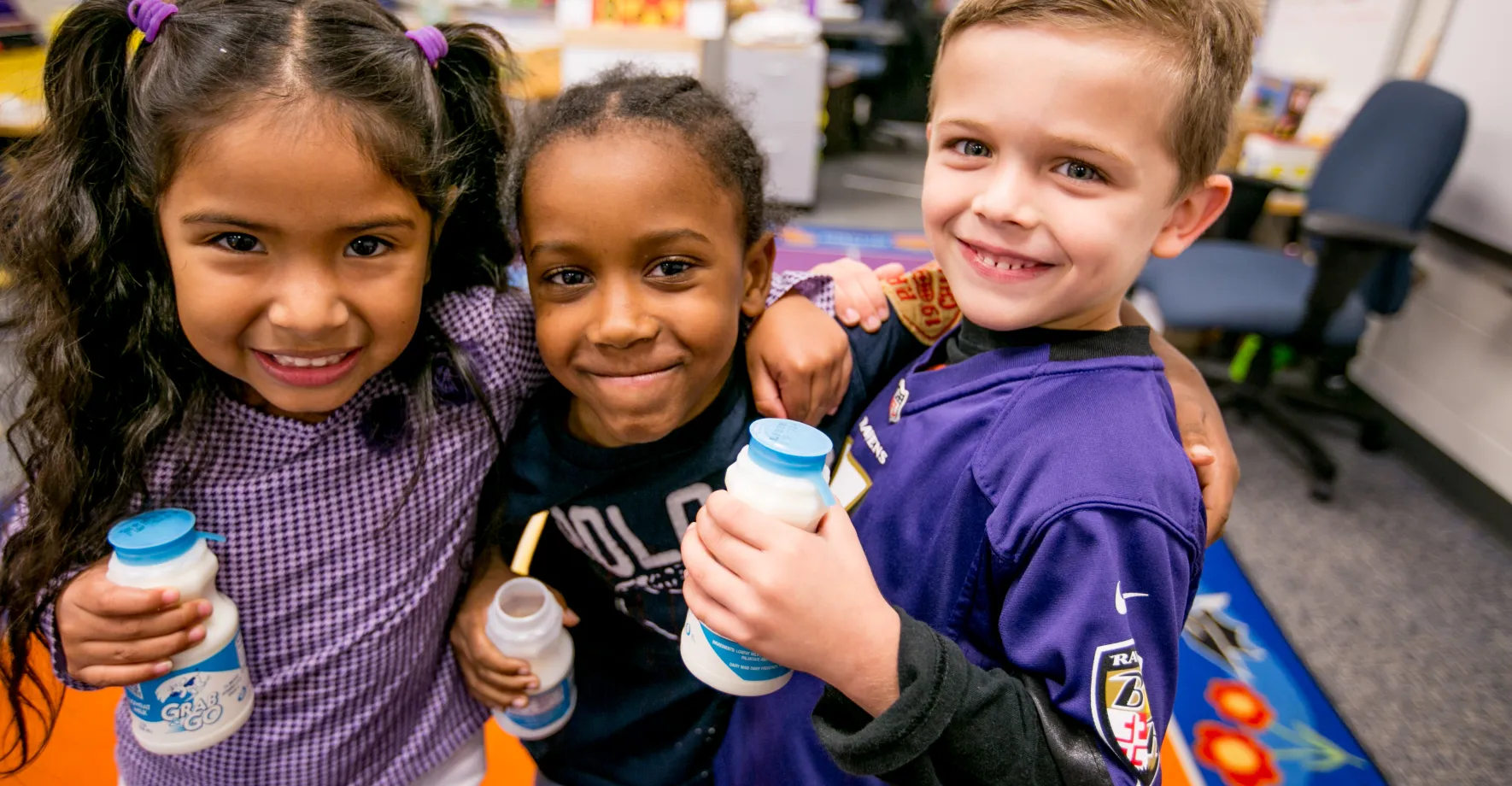 Three elementary school students--a Latina girl with pigtails, a Black boy with cornrows, and a white boy with blond hair--smiling while holding bottles of milk during school breakfast.