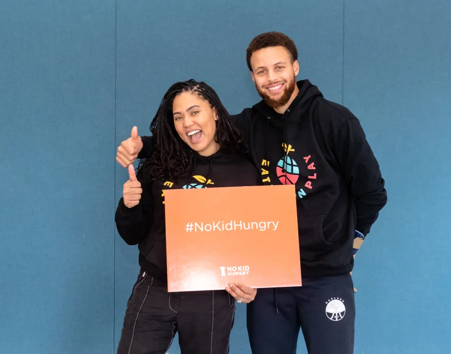 Steph and Ayesha Curry hold an orange No Kid Hungry sign.