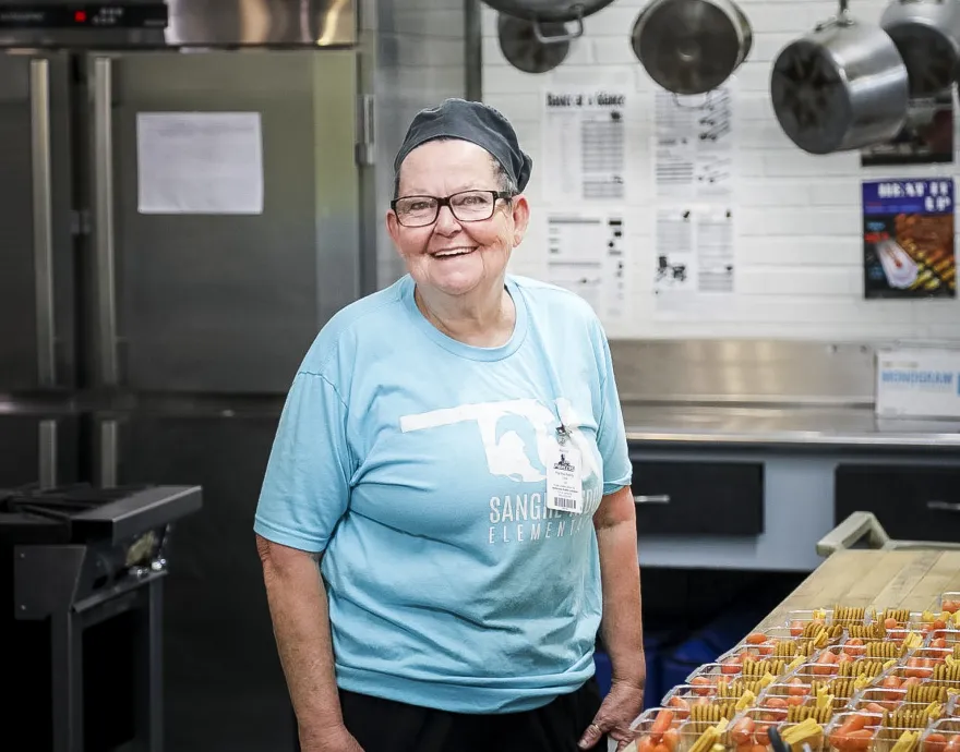 Marty, a cafeteria worker, smiles as she pauses from packing crackers, strawberries and cheese for kids.