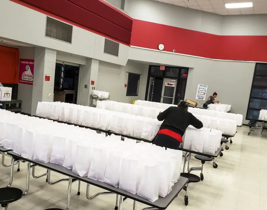 School staff pack rows of lunch sacks in their cafeteria to deliver to kids while school is closed.