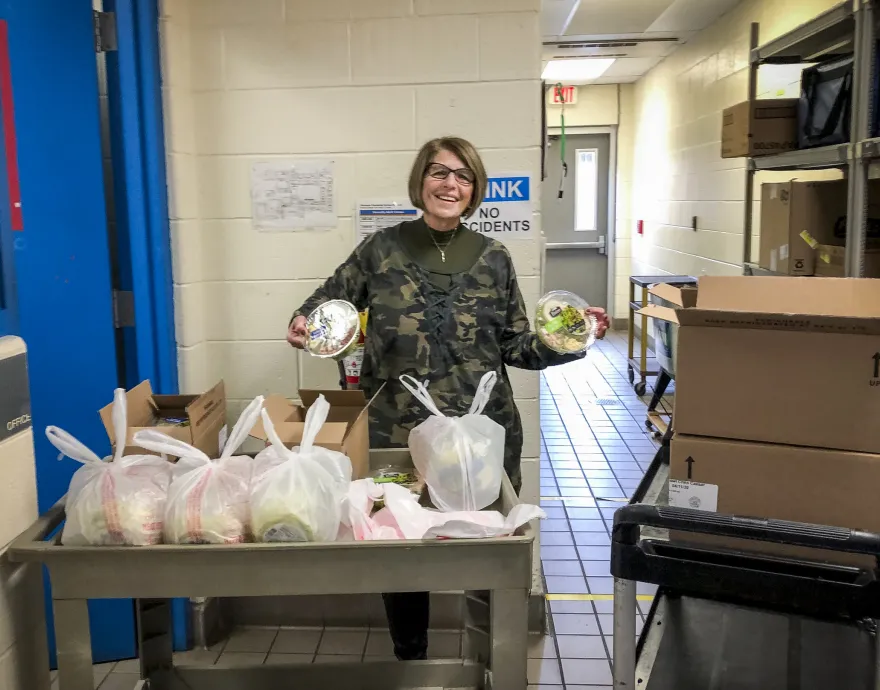 A woman holds up two donated, packaged salads behind a cart packed with meals for families need.