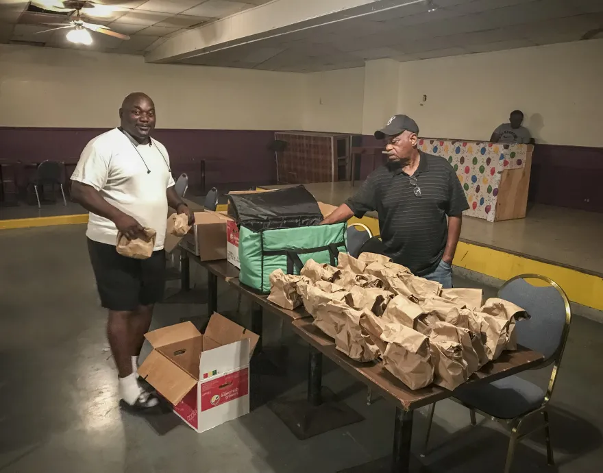 Two men pack paper meals bags at a table in a low-lit room.