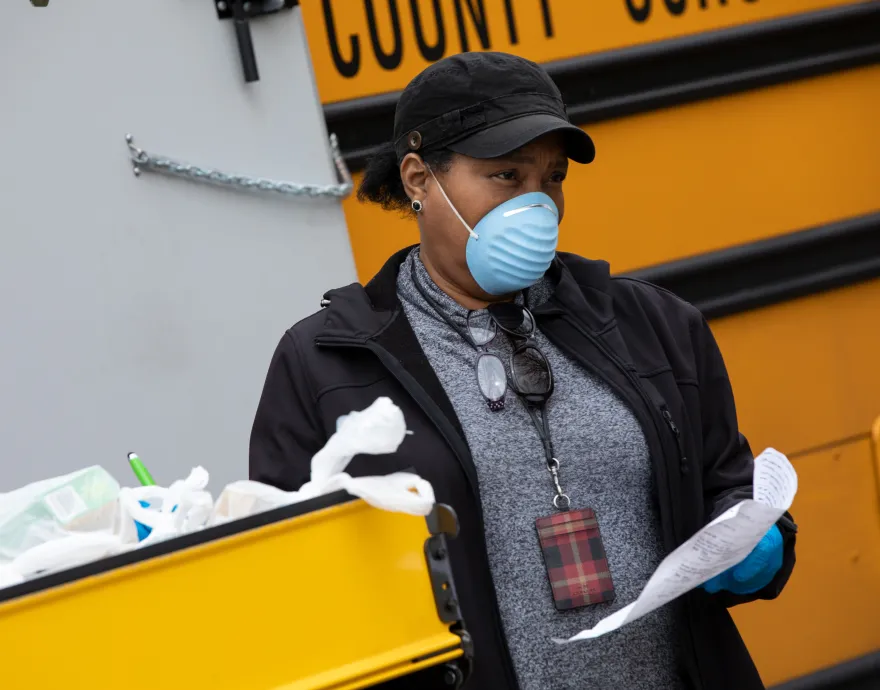 A woman in a mask stands by a school bus and cart of food bags.