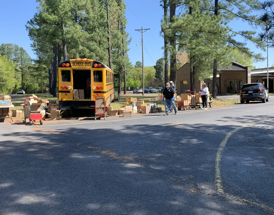 Volunteers in masks pack school buses with sacks of food alongside a wooded road.