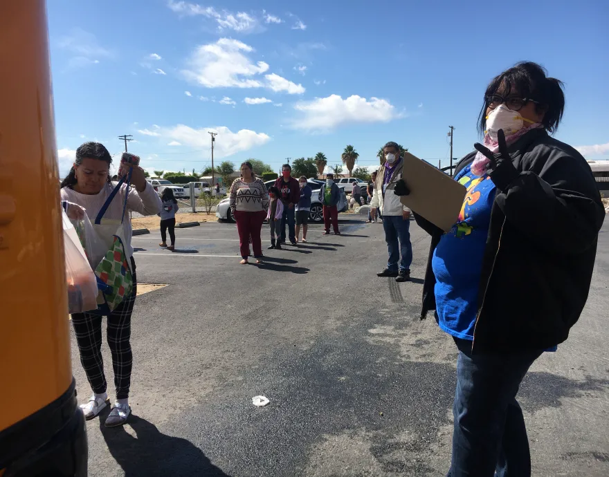 A woman in a mask oversees a line of people waiting for food in a parking lot.