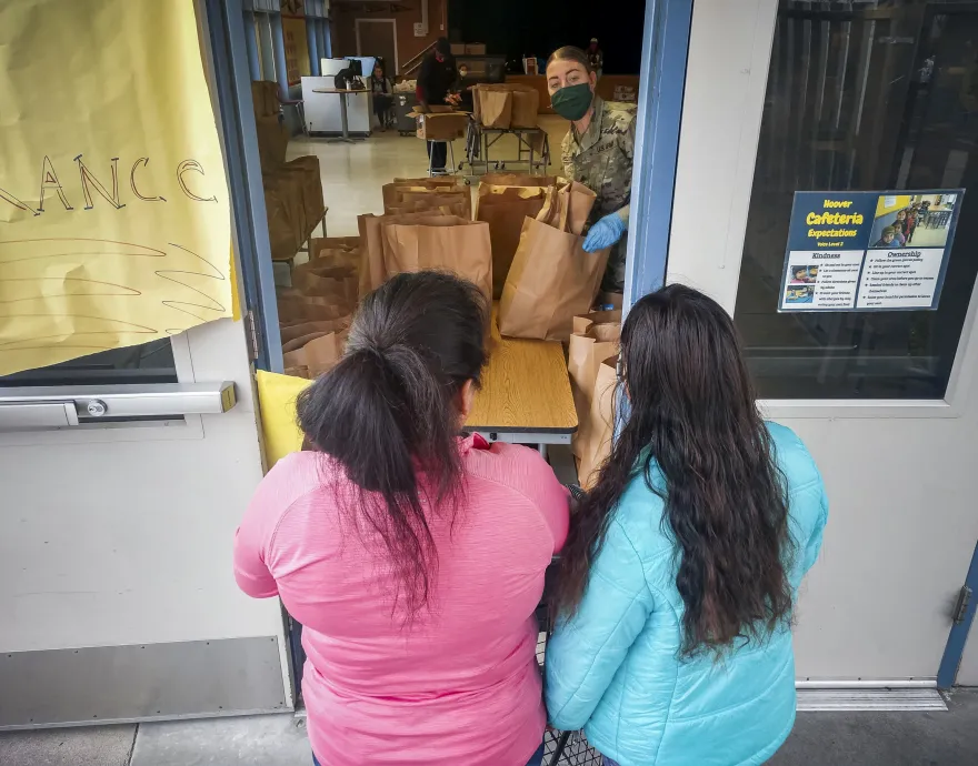 A woman in a mask and military fatigues serves paper bags full of food through a window to two women.