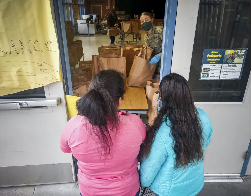 A woman in a mask serves paper bags full of food to two women through a window.