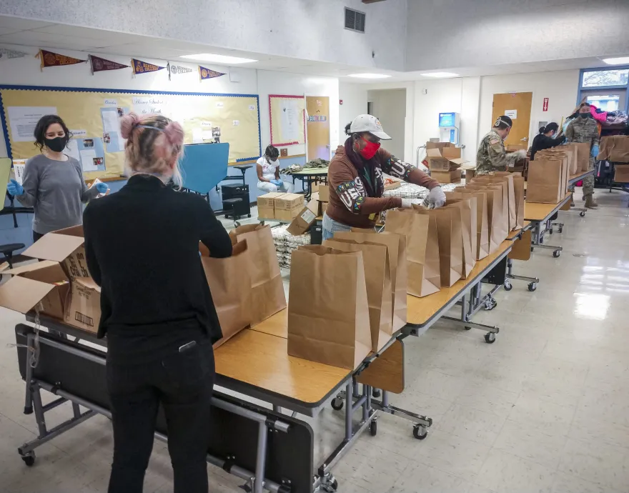 A group packs meals in paper bags on tables in a cafeteria.