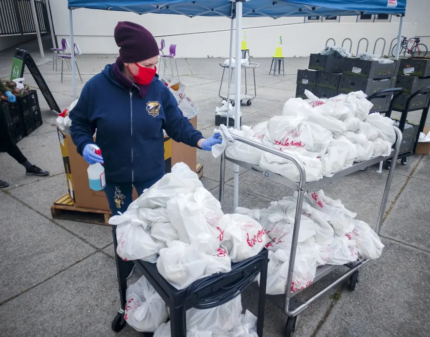 A woman wearing a mask stands by a cart full of plastic bags of meals.