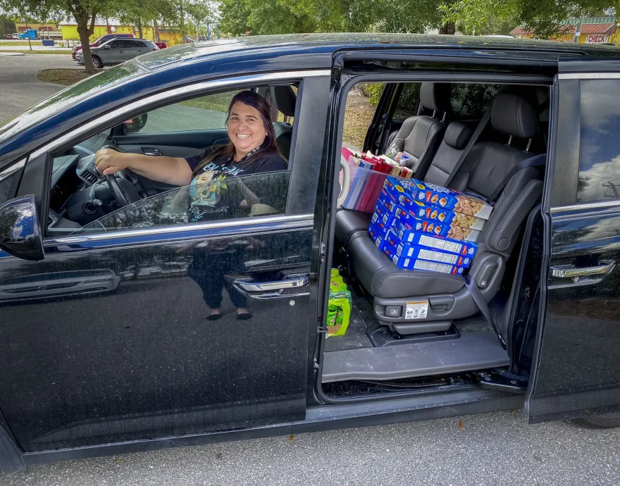 A woman smiles behind the steering wheel of a van loaded with food.