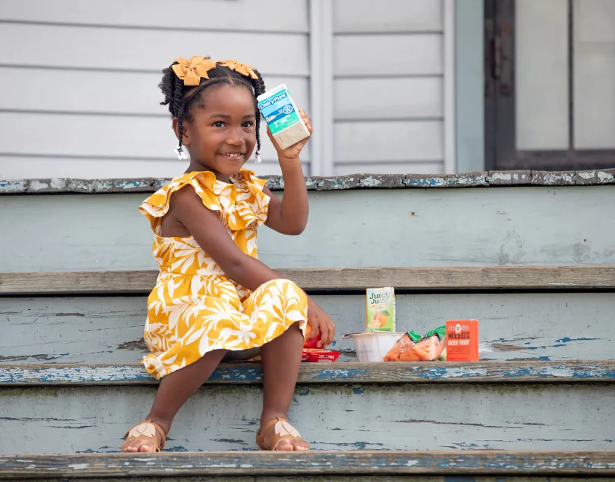 Little girl on a porch holding a box of milk. She is wearing a yellow dress and is very cute