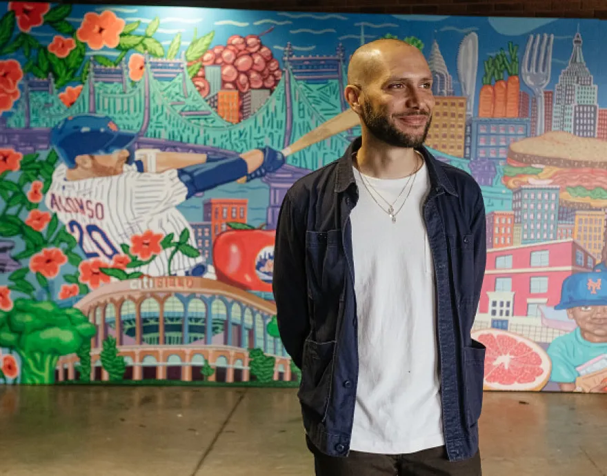 The artist stands in front of his mural, a painted baseball player swinging in the background.