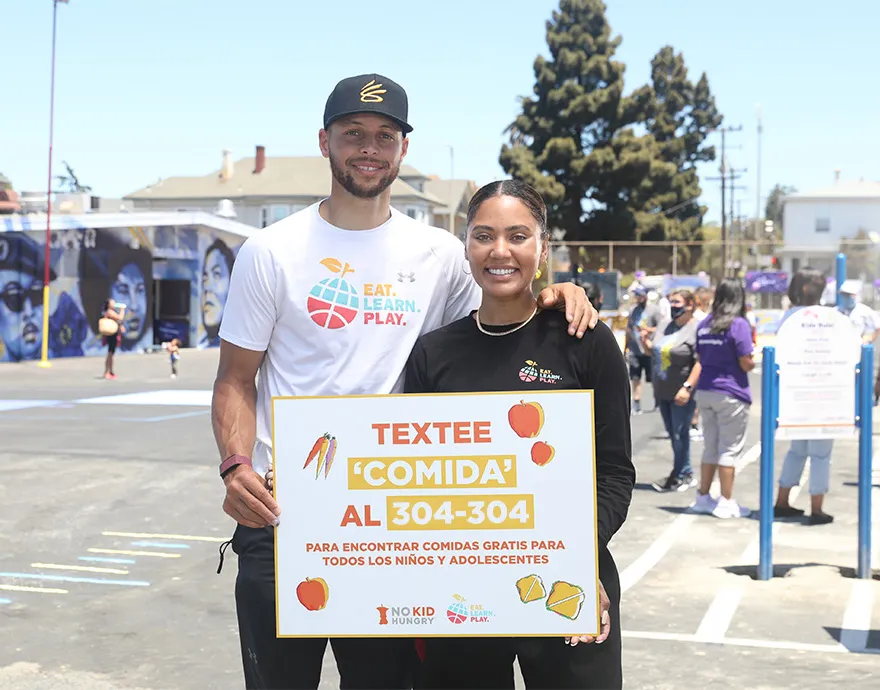 Ayesha and Stephen Curry hold a sign promoting our summer meals texting line in Spanish.