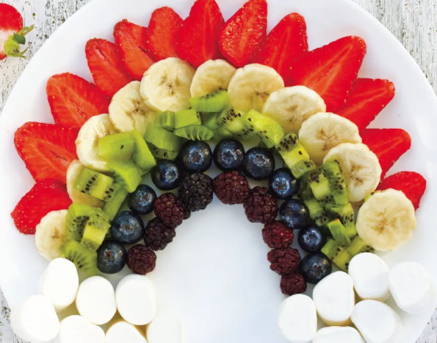 Sliced fruit, berries, and mini marshmallows arranged on a plate to form a rainbow and clouds