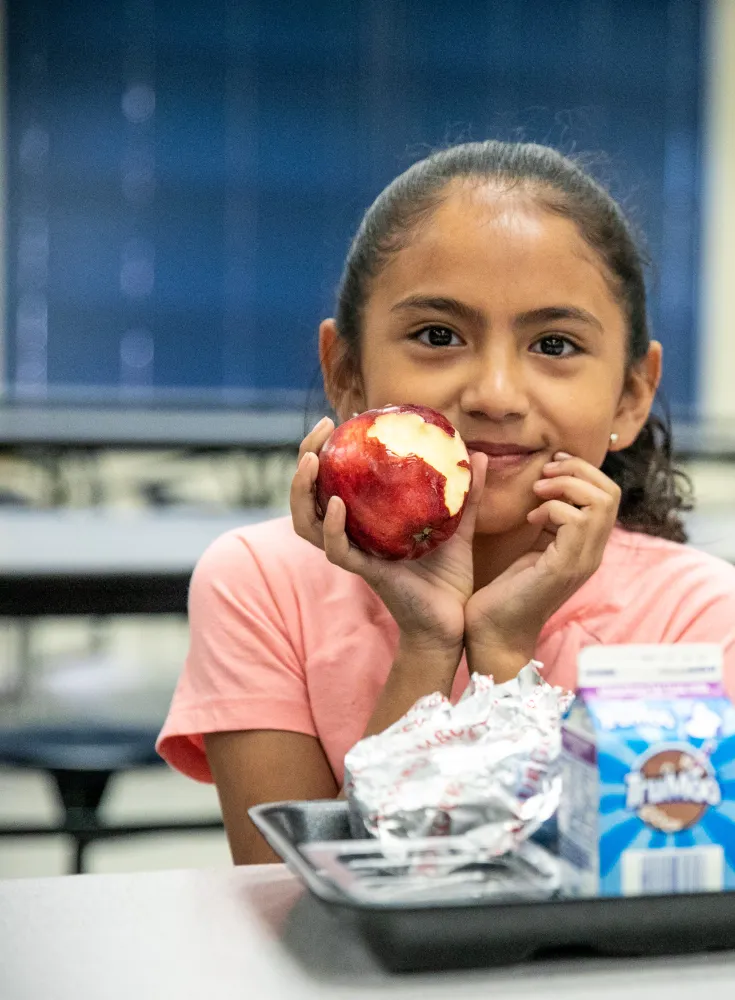 Latino girl eating apple in from of lunch tray