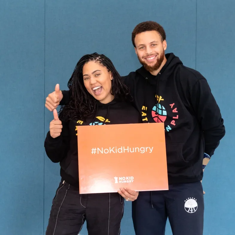 Steph and Ayesha Curry hold an orange No Kid Hungry sign.