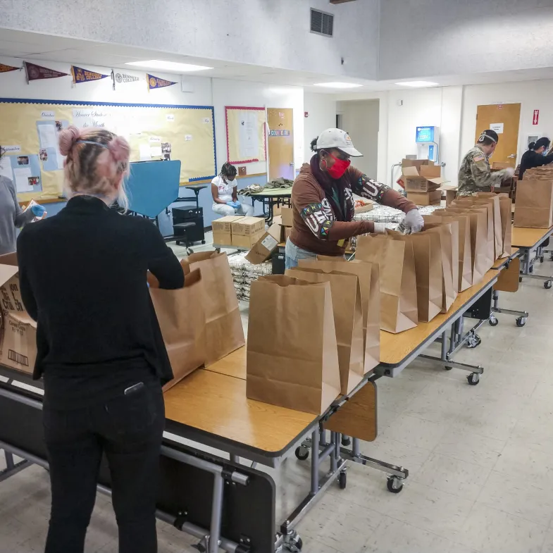 A group packs meals in paper bags on tables in a cafeteria.