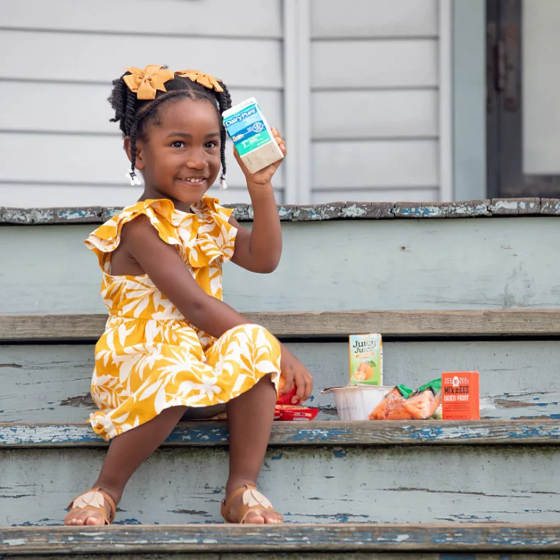 Little girl on a porch holding a box of milk. She is wearing a yellow dress and is very cute