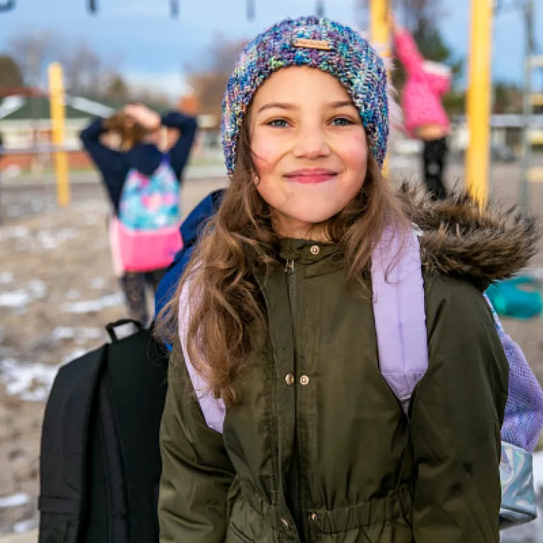 A girl in a beanie and coat smiles on her school playground.