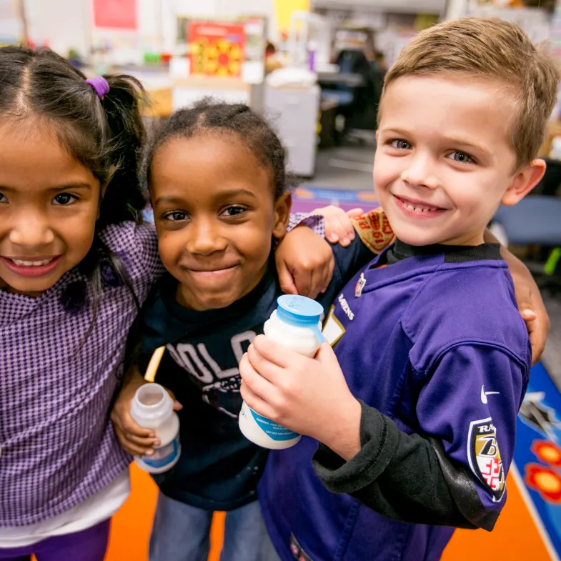 Three elementary school students--a Latina girl with pigtails, a Black boy with cornrows, and a white boy with blond hair--smiling while holding bottles of milk during school breakfast.