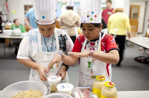 Two kids wearing aprons and chef hats looking at baking ingredients