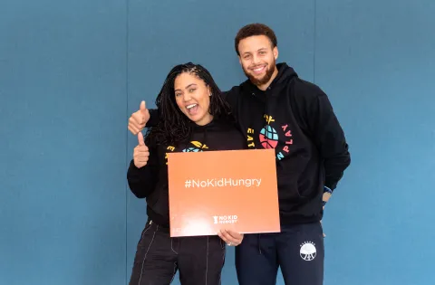 Steph and Ayesha Curry hold an orange No Kid Hungry sign.