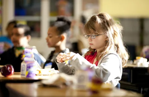 A little girl with glasses eats breakfast in her classroom.