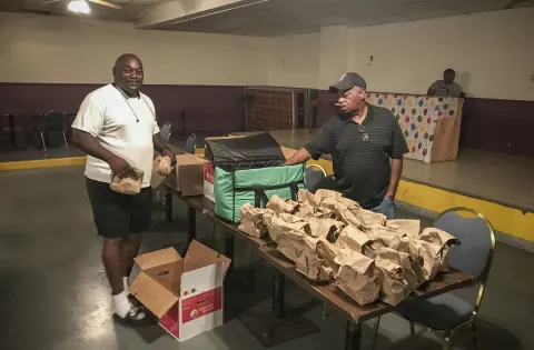 Two men pack paper meals bags at a table in a low-lit room.