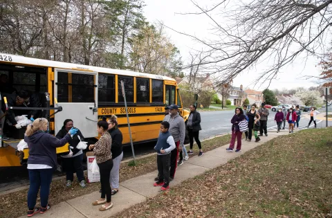 A line of people wait by a school bus for a meal bag.