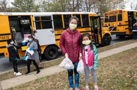 A mother and daughter wear face masks and carry their meals away from the school bus.