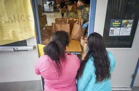 A woman in a mask and military fatigues serves paper bags full of food through a window to two women.