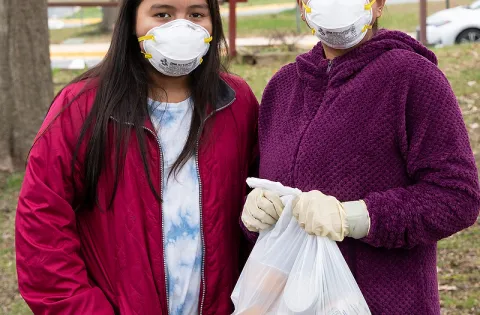 A woman and a girl wearing masks and holding bags of food outside of an elementary school.