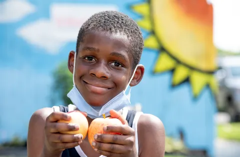 Black kid with mask under the chin holding two apples and happy